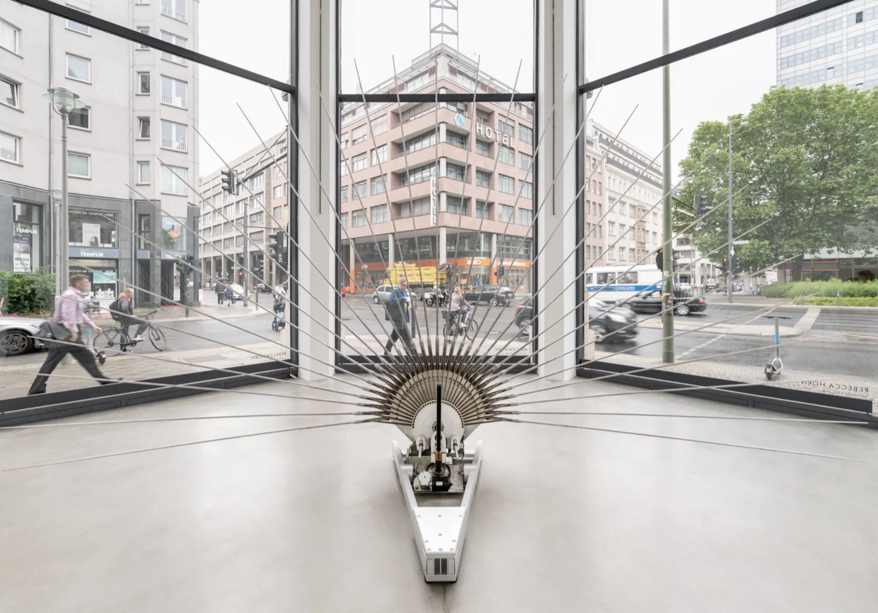 Fächerförmige Metallskulptur in Galerie vor großen Fenstern, Stadtverkehr und Gebäude draußen sichtbar.